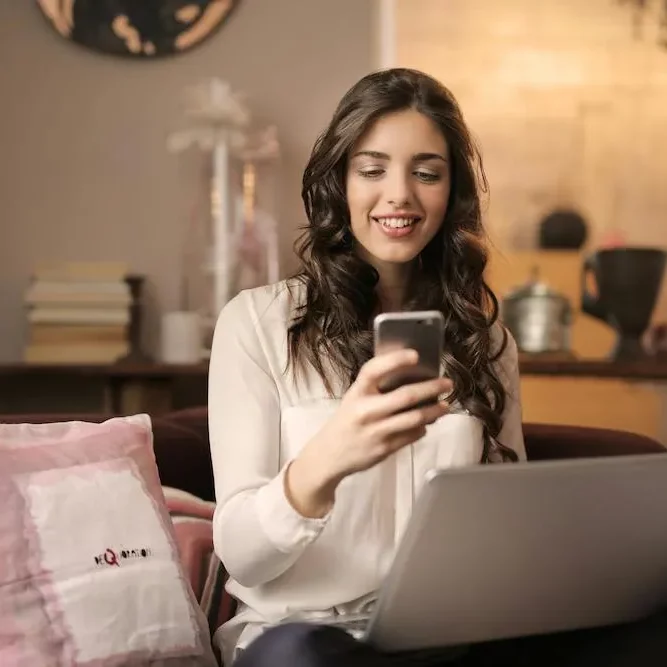 smiling woman looking at phone with laptop on her lap